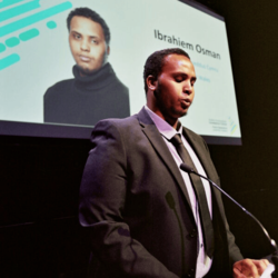 Ibrahiem in black tie, white shirt and black blazer public speaking on stage